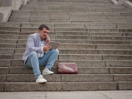 Employee with tablet and talking on a phone, sitting on the urban background. Technology progress concept. Copy space.の写真素材