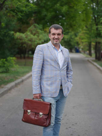 Smiling man walking on a blurred park background. Happy business employee with a briefcase. Financial stability concept.の写真素材