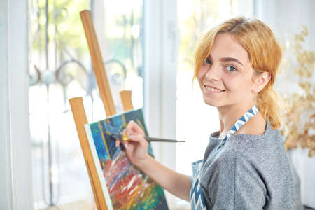 Young smiling girl paints on canvas with oil colors in own workshop. Window on the background. Art...