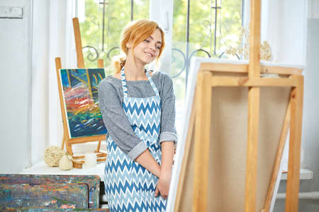 Young smiling girl paints on canvas with oil colors in own workshop. Window on the background. Art...