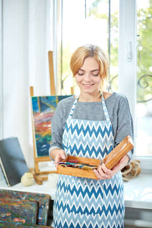 Young smiling girl paints on canvas with oil colors in own workshop. Window on the background. Art concept.の写真素材