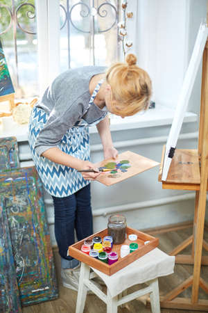 Young smiling girl paints on canvas with oil colors in own workshop. Window on the background. Art...