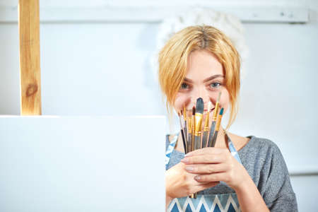 Young smiling girl paints on canvas with oil colors in own workshop. Window on the background. Art...