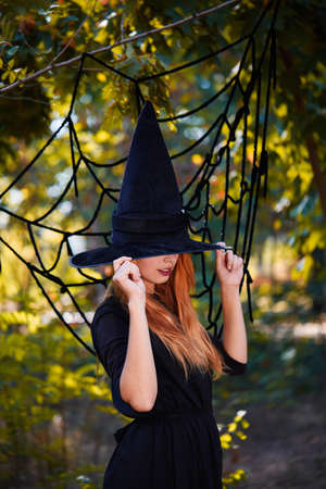 Witch hiding behind black hat. Mysterious ginger witch girl on a forest background. Halloween...