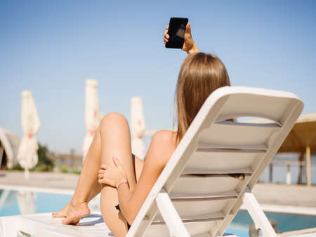 Sexy young girl taking selfies on a chaise-longue on a resort background. Summertime concept. Copy...