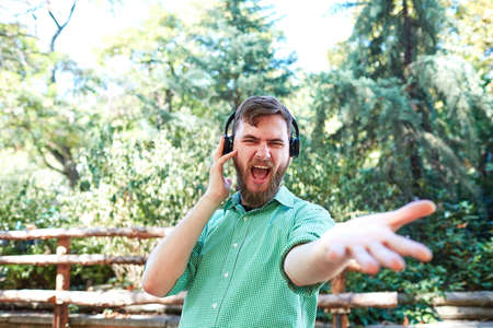 A young guy in a blue shirt listening to music with headphone in the park on nature backround.