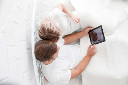 Close-up of a beautiful young couple in a white room using a tablet PC in bed. View from above.