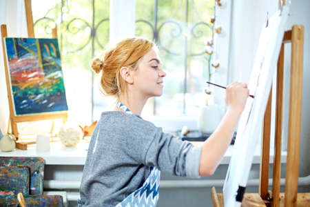 Young smiling girl paints on canvas with oil colors in own workshop. Window on the background. Art concept.の写真素材