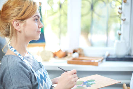 Young smiling girl paints on canvas with oil colors in own workshop. Window on the background. Art...