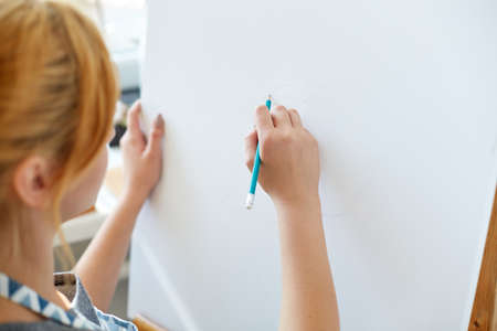 A beautiful artist girl paints on a white canvas with bright multicolored oil paints in a working room. Woman with blonde hair in a blue apron likes to draw. Close-up of hands.の写真素材