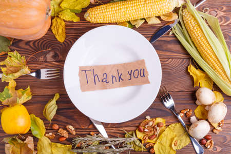 Autumnal concept of vegetables top view of a brown wooden table of corn, mushrooms, pumpkin, bun, leaves with appliances for eating with a note on a plateの写真素材