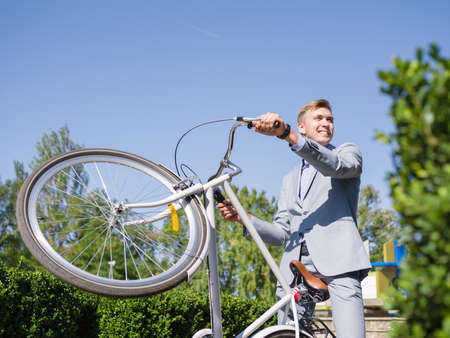 A young man in office attire raises his bicycle in the garden.