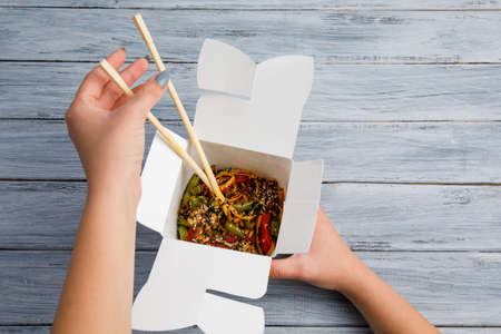 Noodles with chicken and vegetables in a box on a gray table. Female hands and Chinese sticks. The concept of food. Close-up.の写真素材