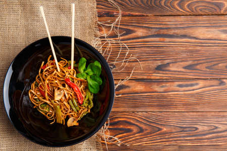 Traditional Chinese dish on a round black plate, rice noodles, cabbage or green cabbage and fried vegetables. Chinese chopsticks. On a wooden brown table. Close-up. View from above.の写真素材