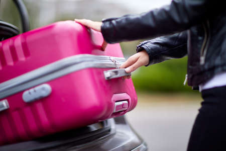 A beautiful girl in a leather jacket picks up a suitcase to put in the trunk of a black carの写真素材