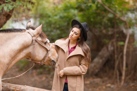 Beautiful young woman in a black hat with a brown horse outdoors.の写真素材