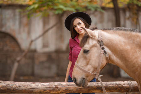 Beautiful young woman in a black hat with a brown horse outdoors.の写真素材