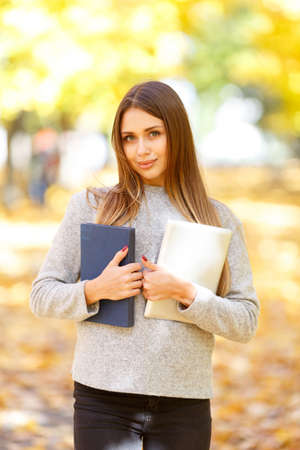 A beautiful girl in a white sweater is holding a book and a tablet on the background of autumn park.の写真素材