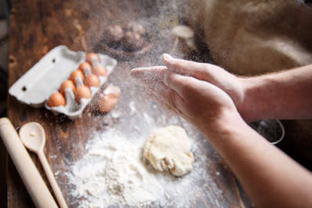 A white man shakes flour from his hands over a wooden table with ingredients. View from above.