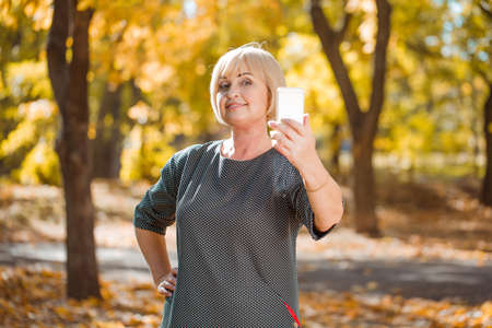 An attractive middle-aged woman walking in an autumn park with gadgets on a blurred background.