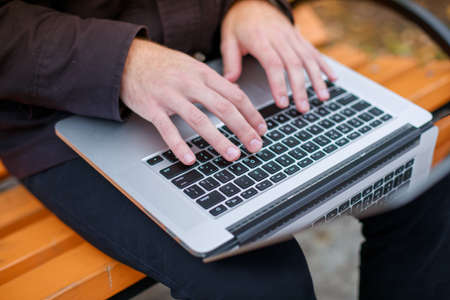 Close-up of a man's hand typing on a laptop in a park on a bench. View from above.