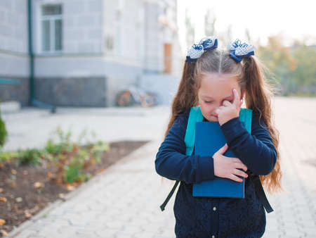A cute little girl with a backpack and a book in her hands is standing outside.