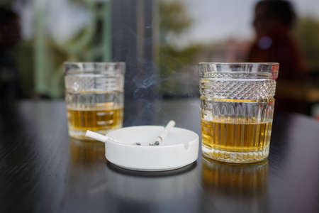 A top view on a two glasses of beer on the dark wooden table. A white ashtray with buutts on the dark background. Close-up of objects. Drink concept.の写真素材
