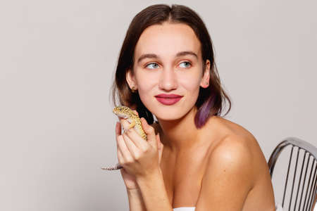 A beautiful girl bare shoulders sits on a chair and holds an iguana gecko in her hand and presses her to her face.の写真素材
