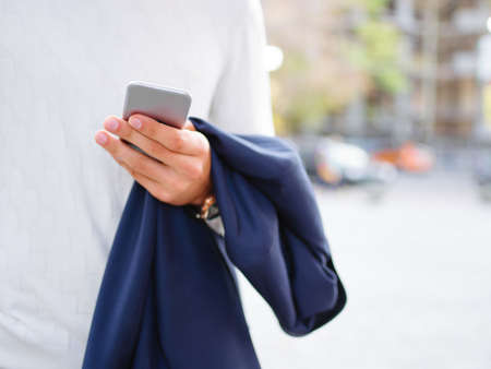 A man in white golf stands on the street holding a mobile phone and a jacket. Outside. Close-up.の写真素材
