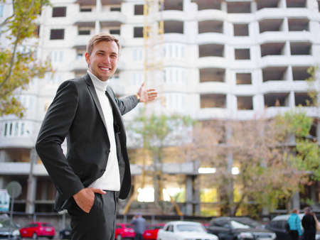 A young guy in a black suit is standing on the street and showing to an unfinished building. Outside.の写真素材