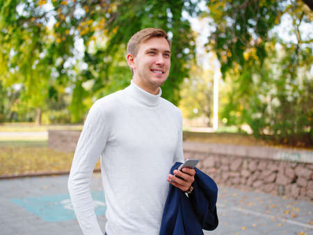 A man stands on the street, looking into the distance and holding a mobile phone and a jacket. Outside.の写真素材