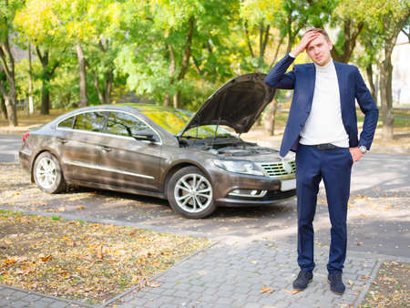 A frustrated guy stands on the street near a broken car and holds his head. Outside.