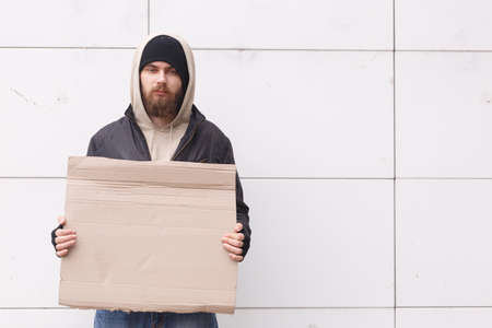 A homeless man stands near a wall in cold weather with a cardboard sign. Outdoor.の写真素材