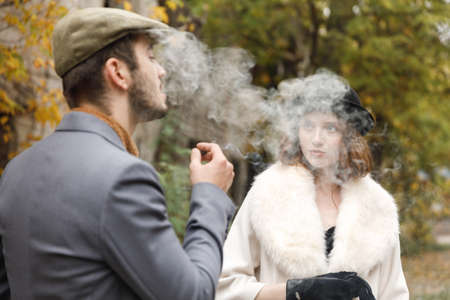 A retro crime couple walking outdoors. The guy smokes a cigar and starts up cigar smoke on the girl. Outdoors.の写真素材