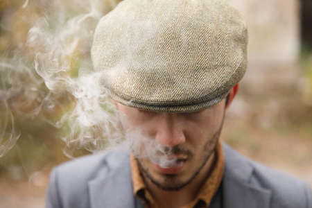 A crime man with a stubble and in a hat smoking a cigar and starting a smoke. Retro. Outdoors. Close up.の写真素材
