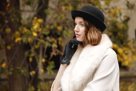 A lady in a hat, gloves and a light coat is looking out, holding her hand by the face. Retro. Outdoors.の写真素材