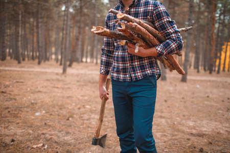 A man in a plaid shirt and blue pants with an ax and firewood at hand close-up in the autumn forestの写真素材