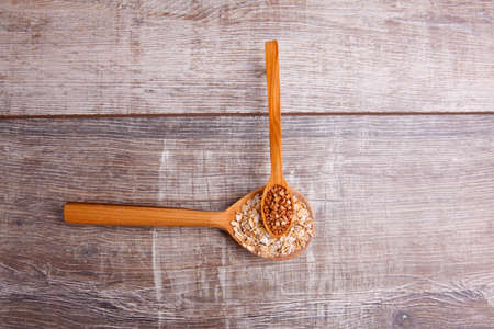 On a gray wooden table in wooden spoons lie buckwheat, and oatmeal. On the spoon with oatmeal lies a small spoon with buckwheat.の写真素材