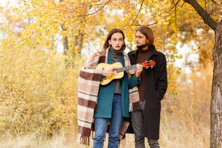 A girl is playing on a ukulele next to a guy on an autumnal blurred backgroundの写真素材
