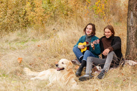 A girl with a ukulele and a boy smiling sitting on a blanket in the autumn forestの写真素材