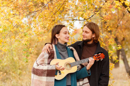 A girl with a ukulele in her hands looks at the guy next to an autumn background