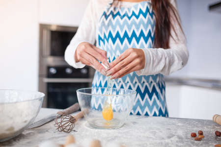 A young girl in the kitchen in a blue apron breaks the egg into a transparent plate. The yolk falls out. On the table, whisk, flour, eggs in a boxの写真素材