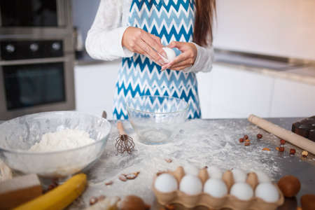 A young woman in an apron smashes eggs in the kitchenの写真素材
