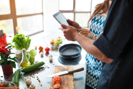 Beautiful young couple uses a digital tablet while cooking food in the kitchen at homeの写真素材