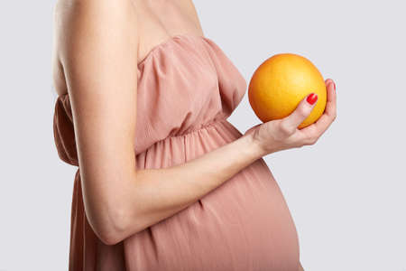 A pregnant girl in a beige dress stands sideways and holds an orange. Side view. Studio, white background.の写真素材