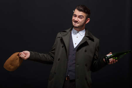 A man on a black background holds a bottle in his hand and a brown cap behind his ear a cigarette.の写真素材