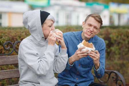 Portrait of a hungry young couple who eat burgers in a park on a bench and smiling.の写真素材