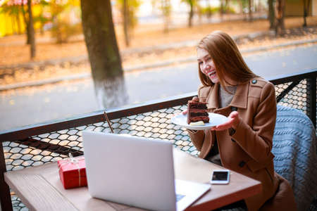 A girl in an outdoor cafe with a laptop talking to someone via a video call, showing her cakeの写真素材