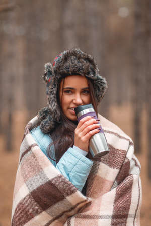 A girl in a warm hat and covered in a blanket, drinks from a hot mug in the forest.の写真素材
