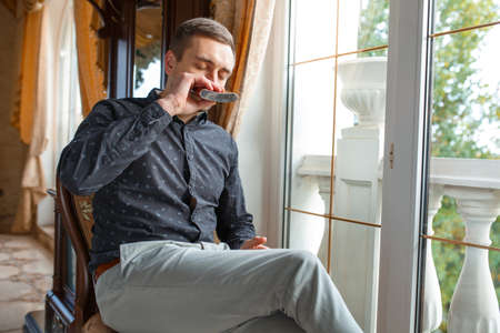 A guy drinks from a jar sitting on a chair by the window